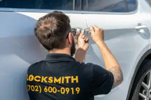 A locksmith is expertly unlocking a silver car door under the bright sun. He wears a black polo shirt with "LOCKSMITH" and a phone number in yellow on the back, embodying his role as one of the best locksmith services. Sporting short hair, a beard, and glasses, he's focused on using his specialized tool.