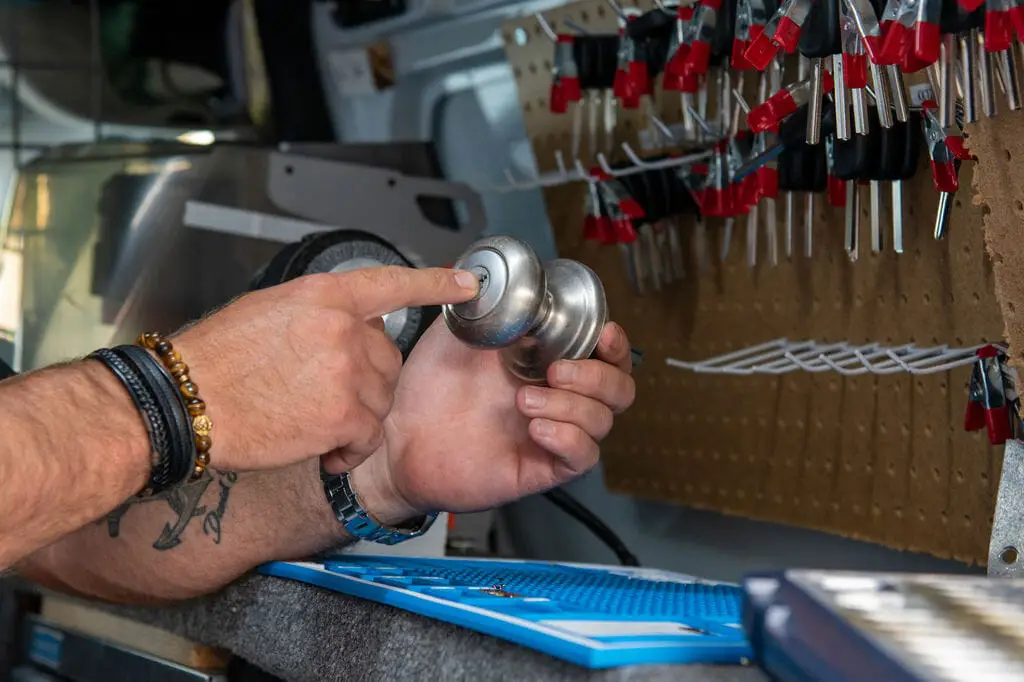 A person with tattooed arms and wearing bracelets holds a metallic door knob, pointing at it. The background reveals a board with numerous locks and keys, alongside a partially visible organized tool set, suggesting the workspace of a locksmith near me.