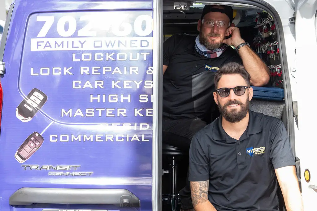 Two men are sitting in the back of a van, which showcases services like rekey, lockout assistance, and car key solutions. A "Family Owned" sign is visible. The man on the left sports a cap and glasses, while the one on the right wears sunglasses. Both are casually dressed in black shirts as they discuss being the best locksmiths around.