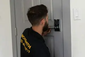 A locksmith from Locksmith Las Vegas, sporting short dark hair and a black shirt with yellow lettering, is installing an electronic keypad lock on a gray door. Focused on securing the device with a screwdriver, he showcases expert locksmith services beside the light switch on the right.