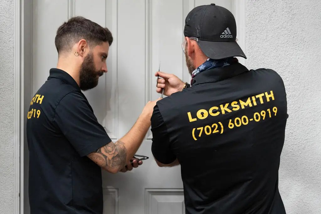 Two locksmiths wearing black shirts with LOCKSMITH (702) 600-0919 on the back work on a door. One man, with tattoos on his left arm, uses tools on the lock. The other, wearing a black cap, assists him. Focused and precise, they offer local locksmith services to unlock the white door on a light gray wall.