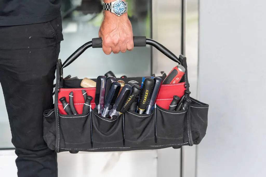 A person wearing a watch stands near a glass door, holding a black and red tool bag filled with various tools essential for locksmith services, including screwdrivers and wrenches. The bag features multiple compartments for securing each tool firmly, its handle gripped tightly.