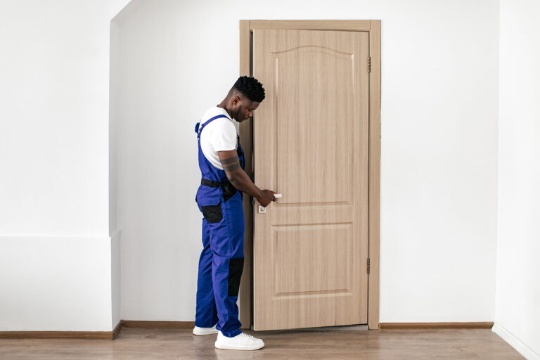 Repair Service. Black Repairman Installing New Entry Door, Fixing Lock And Handle Working Wearing Blue Coverall Uniform In Flat Indoor. Male Renovation Concept.